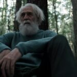 An elderly man with a beard sits thoughtfully in a serene forest setting. Natural and reflective.
