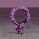 A rusty pink female gender symbol displayed on a textured surface, signifying femininity and resilience.