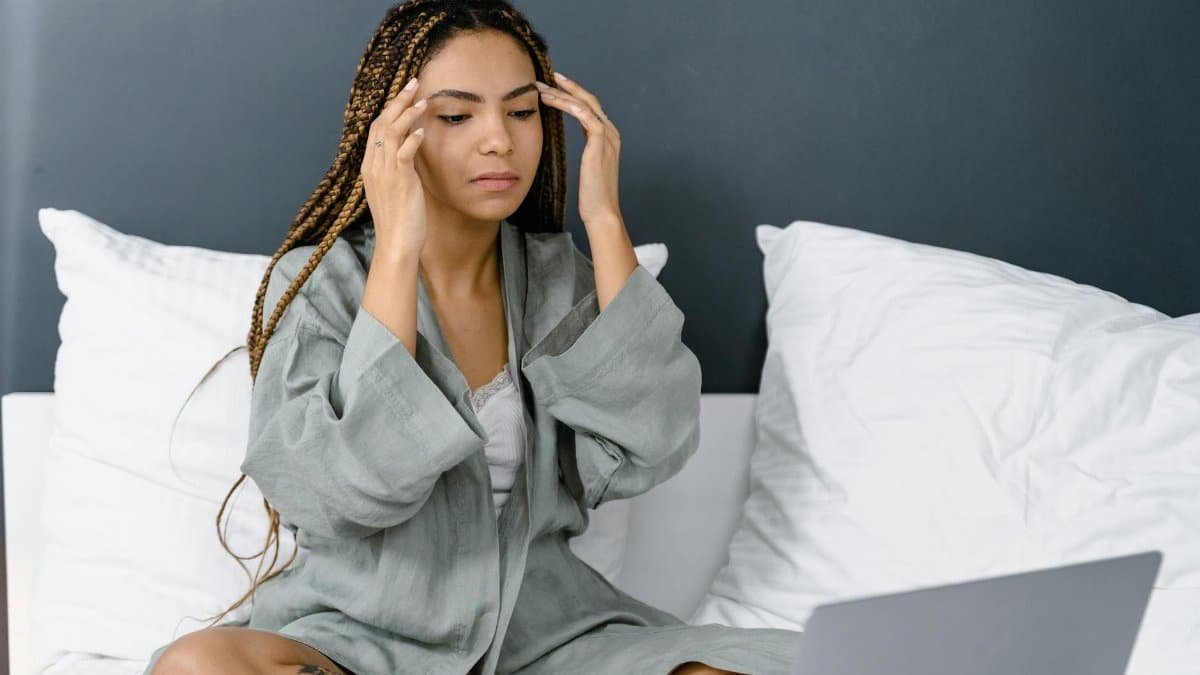 A young woman with braided hair sits in her bedroom, using a laptop, deep in thought.