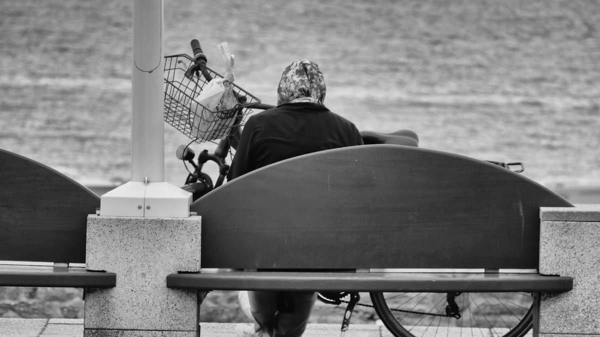 A woman in a headscarf sits on a bench overlooking the sea, with a bicycle nearby, in a peaceful monochrome scene.