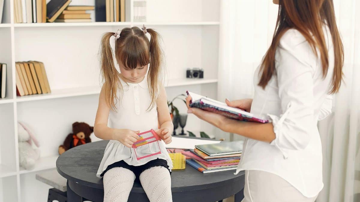 Little child in white dress using abacus for mental arithmetic while sitting on black round table with textbooks and school items against bookshelf during home education with crop young female tutor standing near with diary in hands
