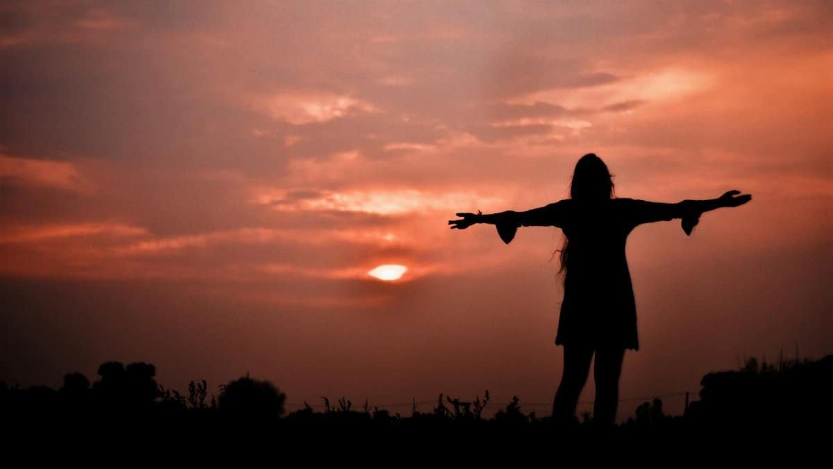 A woman stands in silhouette with arms open, embracing a dramatic sunset in India.