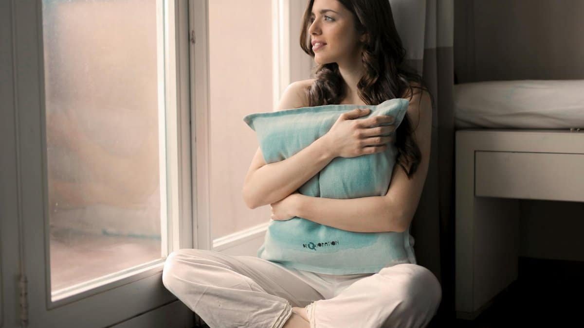 Serene woman sitting indoors, embracing pillow, lost in thought by a bright window.