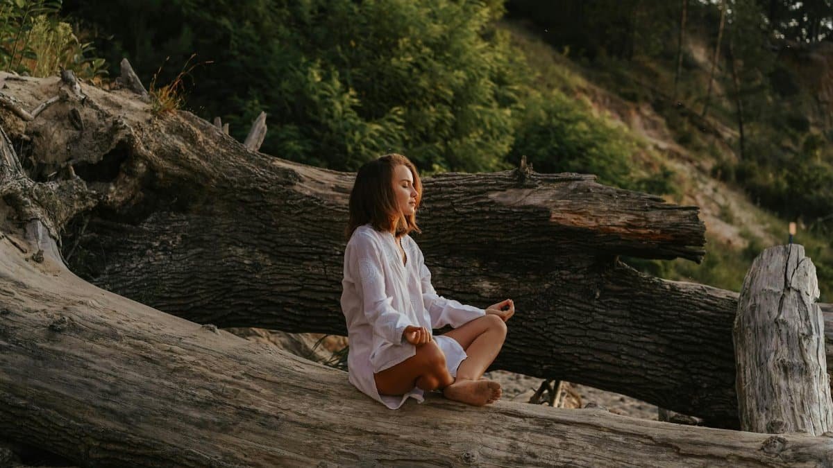 Woman in white shirt meditates on a large log in a forest setting during the day.