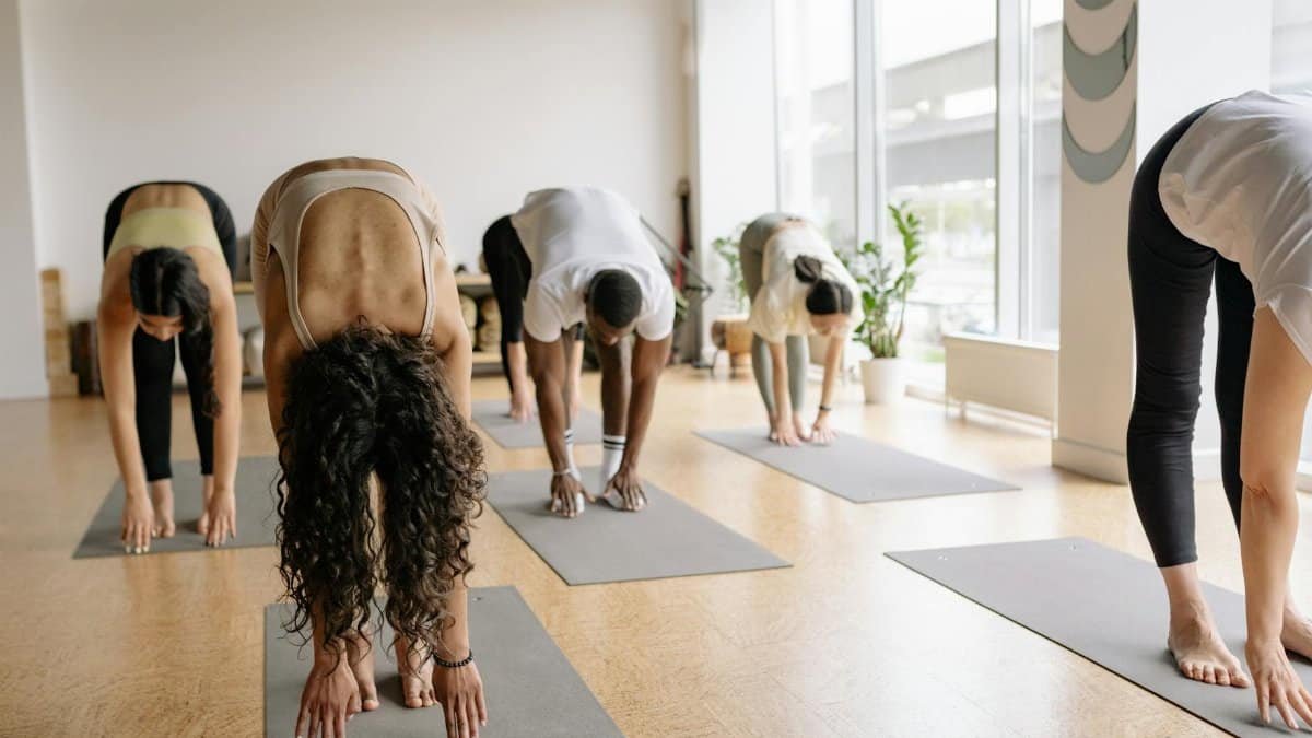 A diverse group performing yoga forward bend pose in a bright yoga studio.