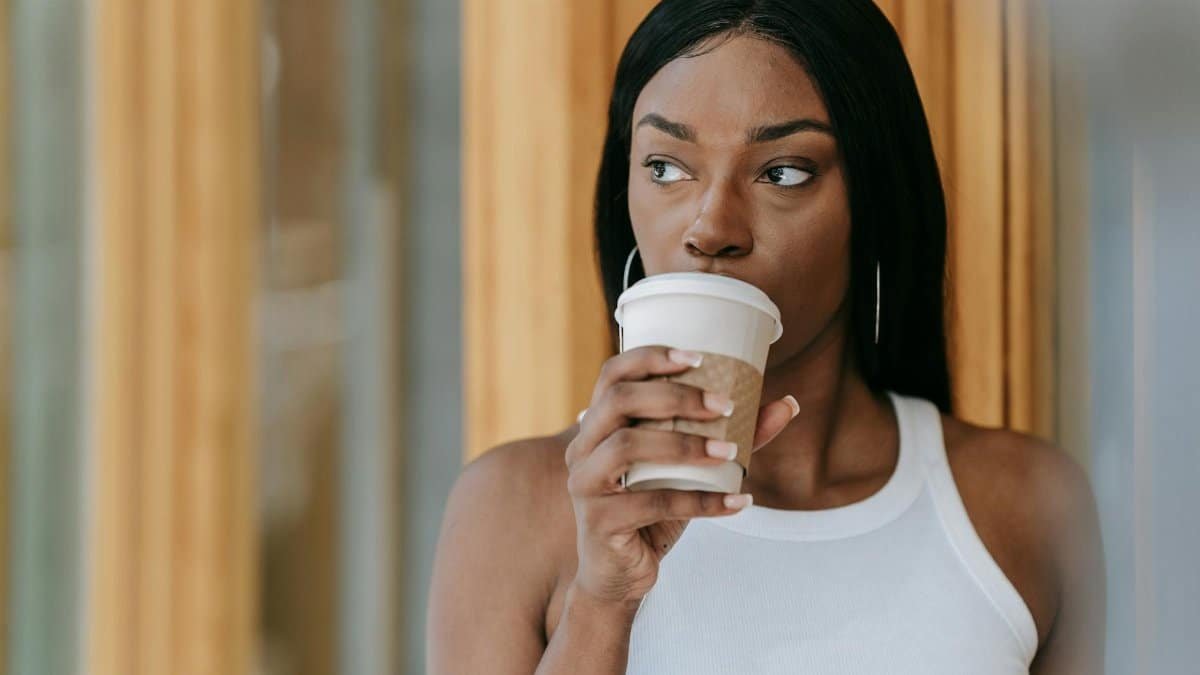 A woman in a white tank top thoughtfully sipping coffee indoors. Warm tones create a serene atmosphere.