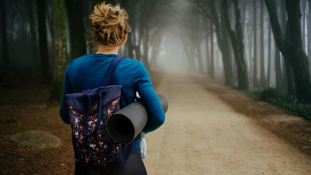 A woman with a backpack and yoga mat walks through a misty forest trail, creating a sense of mystery.