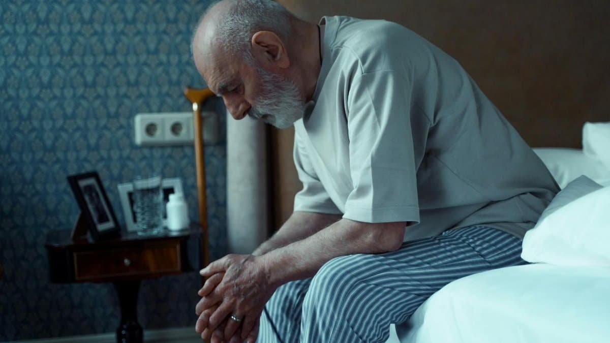 An elderly man sits thoughtfully on a bed, conveying solitude and reflection.
