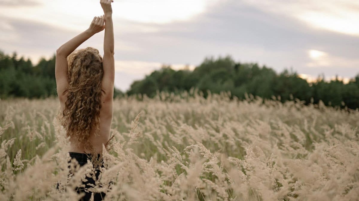 A woman stands with raised arms in a serene field, embracing nature's tranquility.