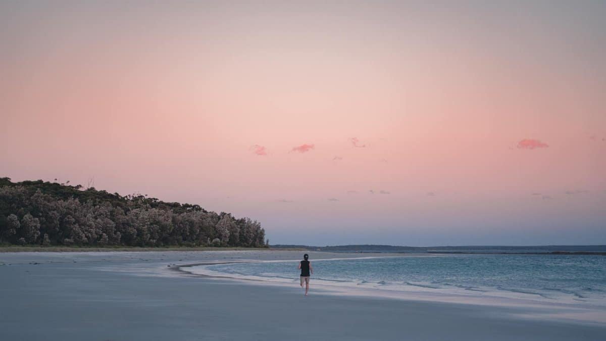 A lone individual walks along a tranquil beach under a pastel dawn sky, offering a sense of peace and solitude.