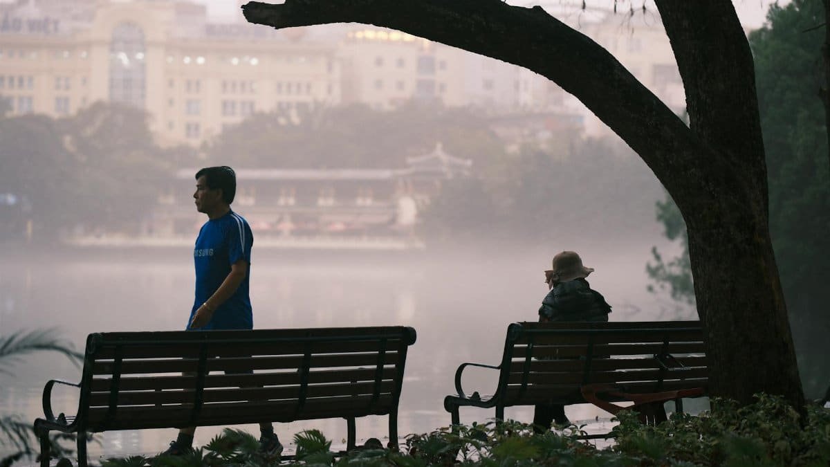 Serene morning scene with a person walking and another on a bench by a misty lake.
