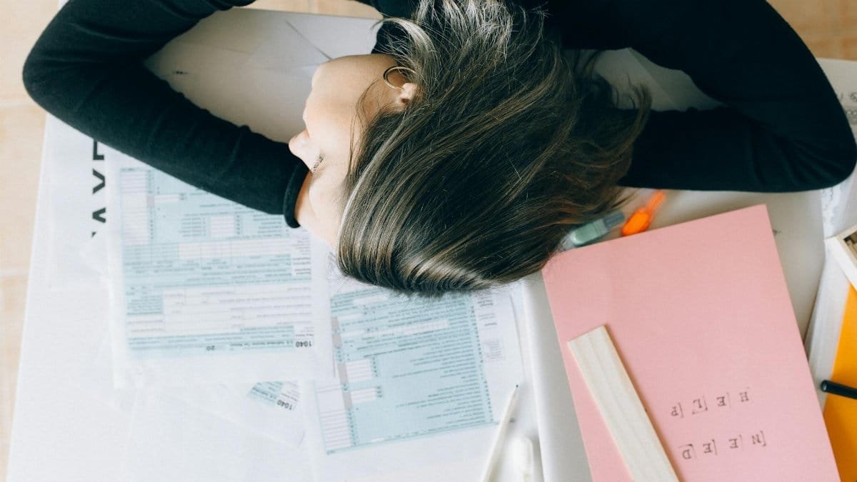 Exhausted woman resting her head on a desk cluttered with papers and a ruler, showing workplace fatigue.