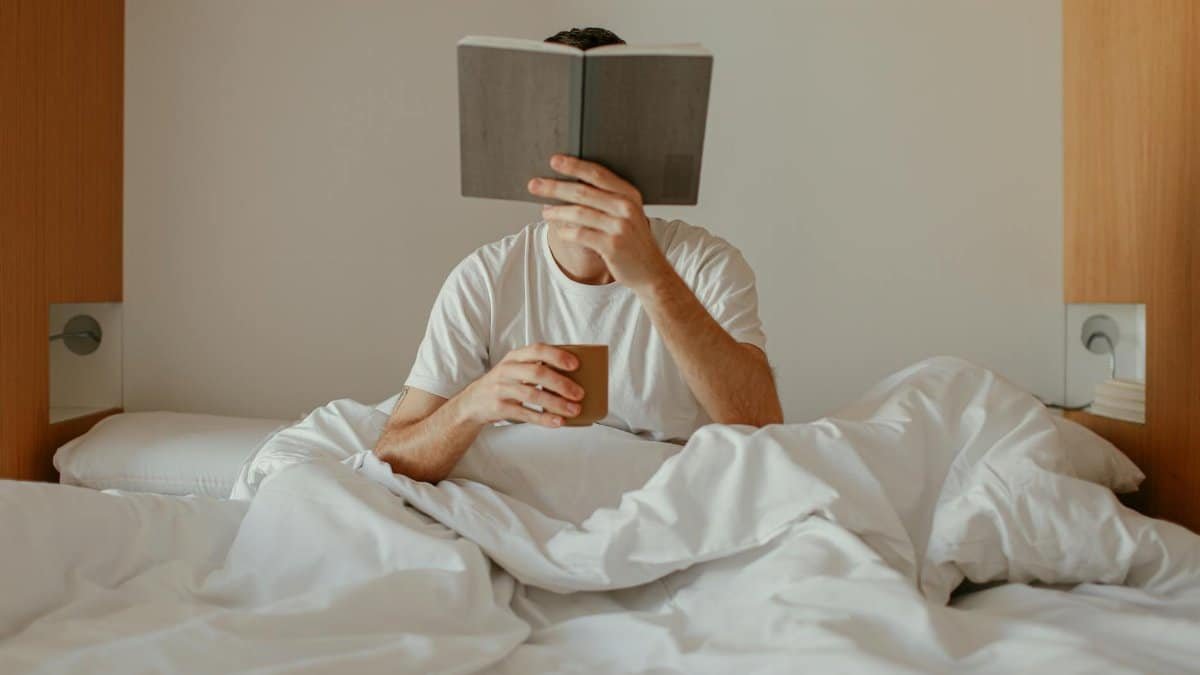 Adult man relaxing and reading a book in bed, holding a cup, cozy morning indoors.