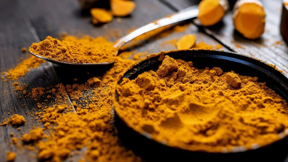 Close-up of vibrant turmeric powder with root on rustic wooden table, emphasizing texture and color.