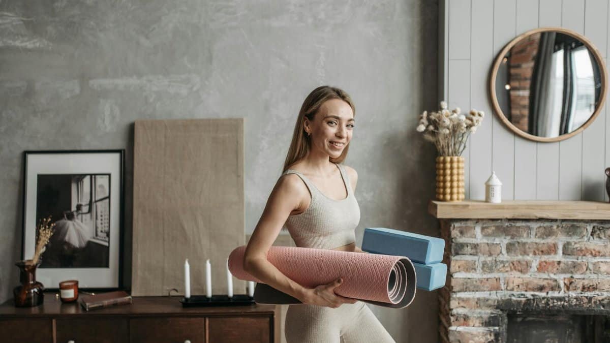 A smiling woman in activewear holding a yoga mat and blocks in a stylish home interior.