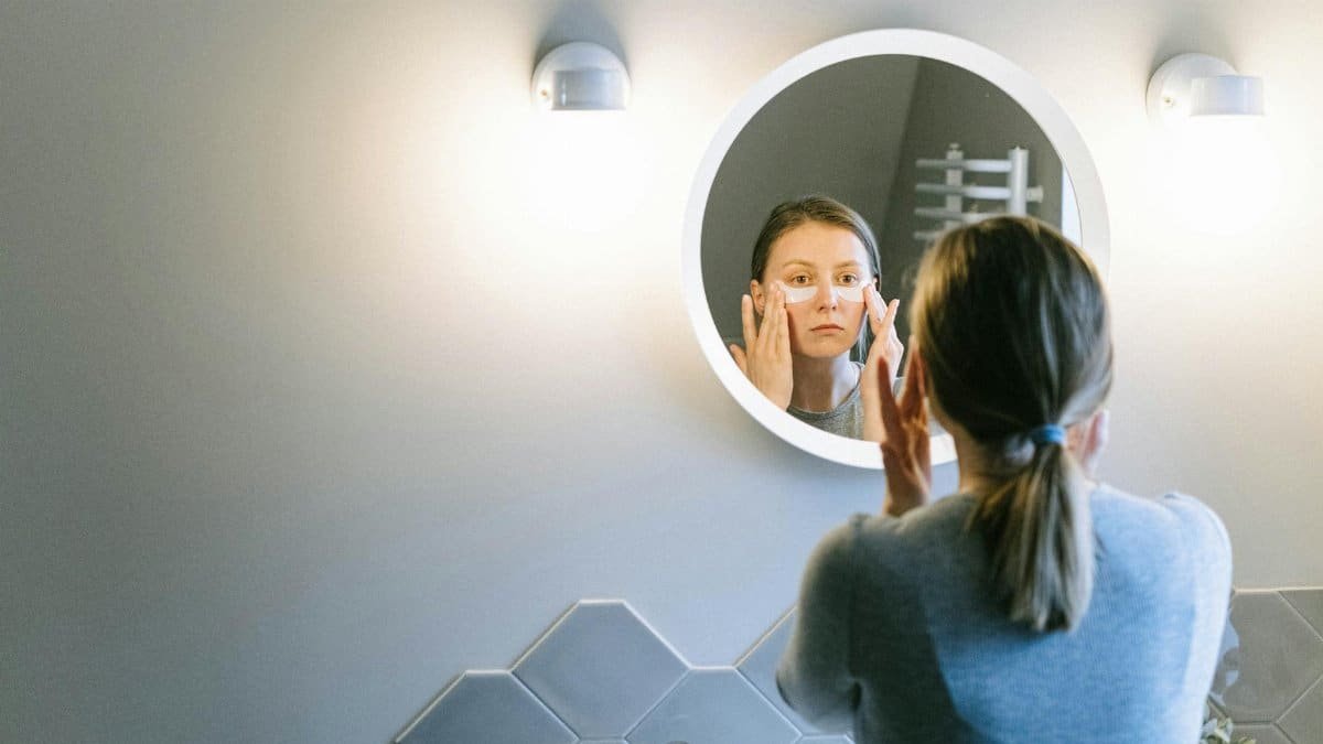 A woman applies under eye patches in a modern bathroom, emphasizing self-care.