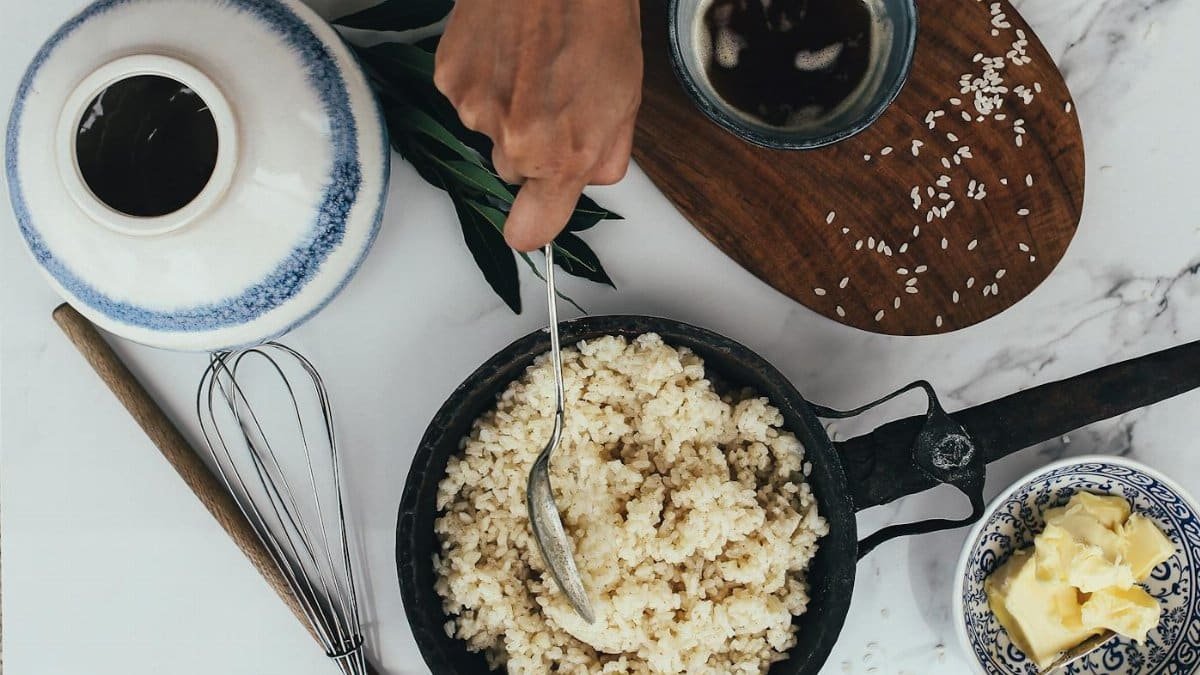 Hand stirring rice in a pan with butter and tea on a marble surface for a homely meal setup.