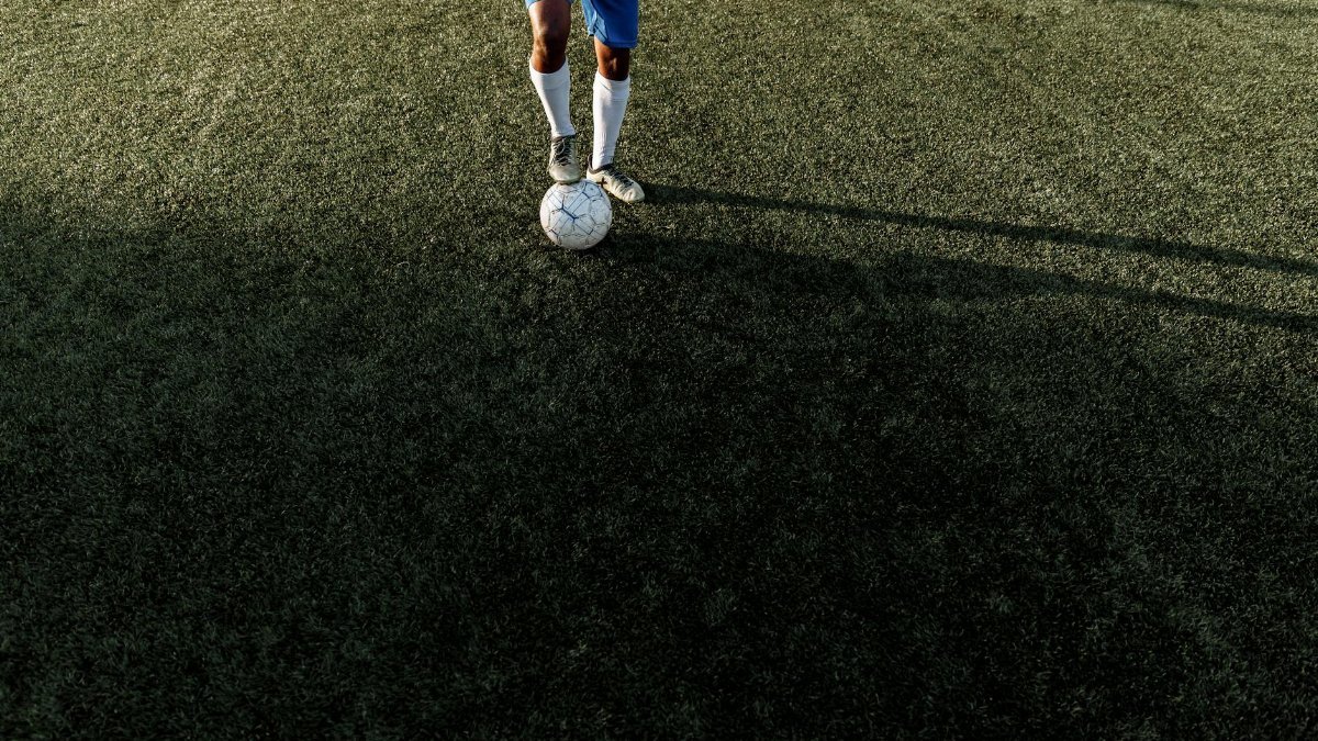 A soccer player stands on a field with a ball under bright sunlight, casting a long shadow.