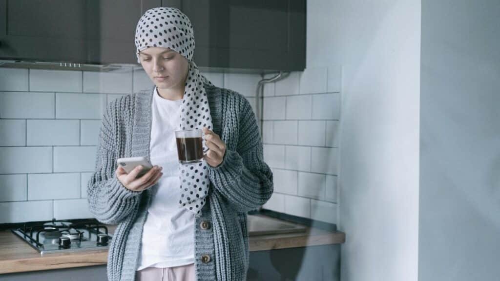 A woman wearing a headscarf and sweater holds a coffee cup and phone in a modern kitchen, symbolizing strength and recovery.