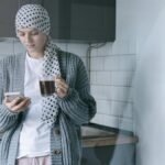 A woman wearing a headscarf and sweater holds a coffee cup and phone in a modern kitchen, symbolizing strength and recovery.