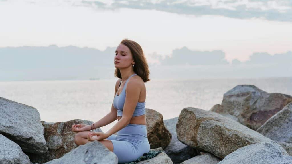 Woman in activewear meditating peacefully on rocks by the ocean at sunrise, promoting wellness and tranquility.