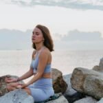 Woman in activewear meditating peacefully on rocks by the ocean at sunrise, promoting wellness and tranquility.