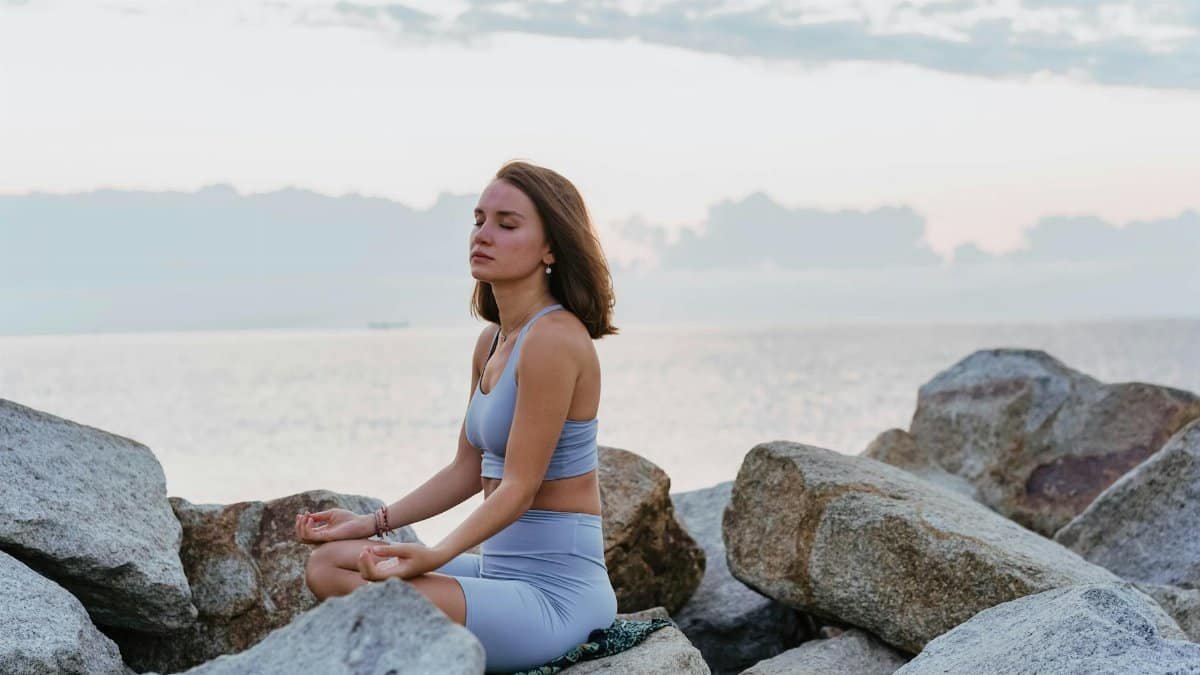 Woman in activewear meditating peacefully on rocks by the ocean at sunrise, promoting wellness and tranquility.