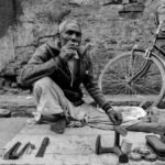 Black and white photo of an elderly cobbler sitting beside a bicycle, showcasing street life.