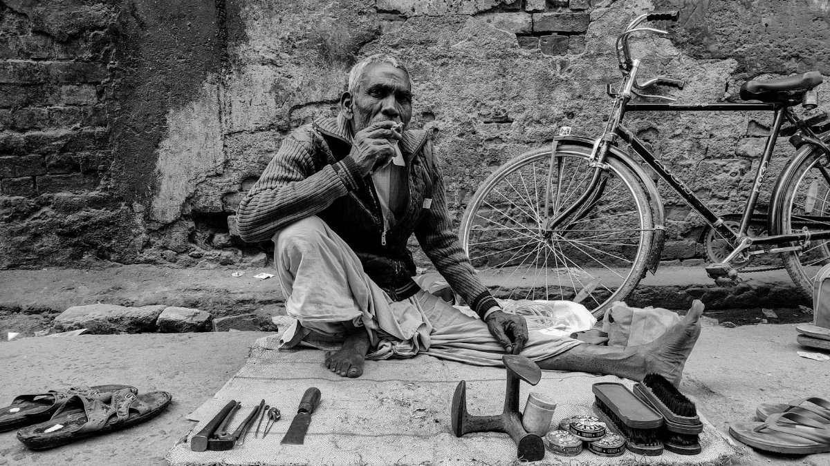 Black and white photo of an elderly cobbler sitting beside a bicycle, showcasing street life.
