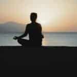 A woman practices meditation with a tranquil ocean sunset backdrop, embodying peace and relaxation.
