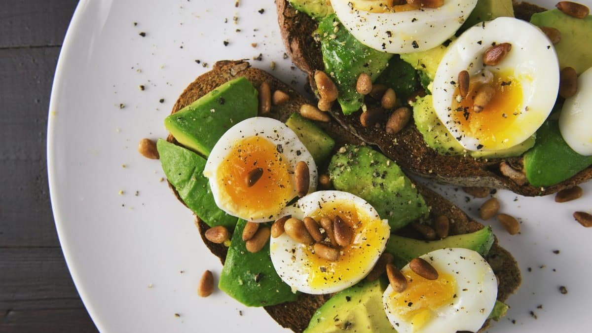 Delicious avocado toast topped with soft-boiled eggs and pine nuts on a white plate.