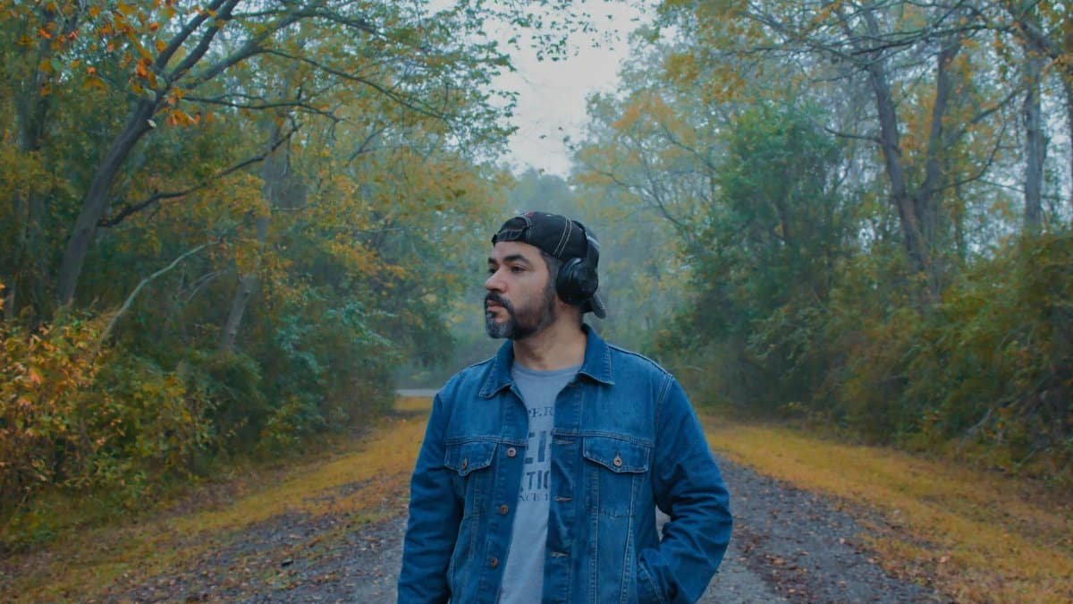 A man in a denim jacket stands in a serene autumn forest, listening to music with headphones.