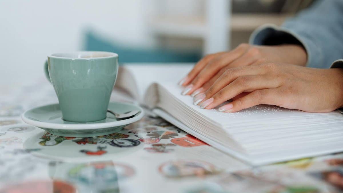 Close-up of a woman's hands reading a braille book while enjoying coffee, emphasizing sensory engagement.