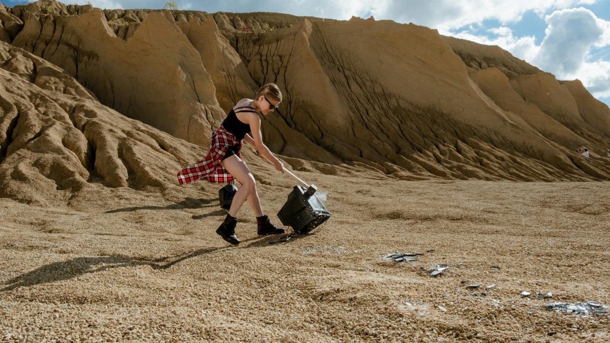 A young woman in a rocky desert scene breaks a vintage TV with a sledgehammer under a cloudy sky.