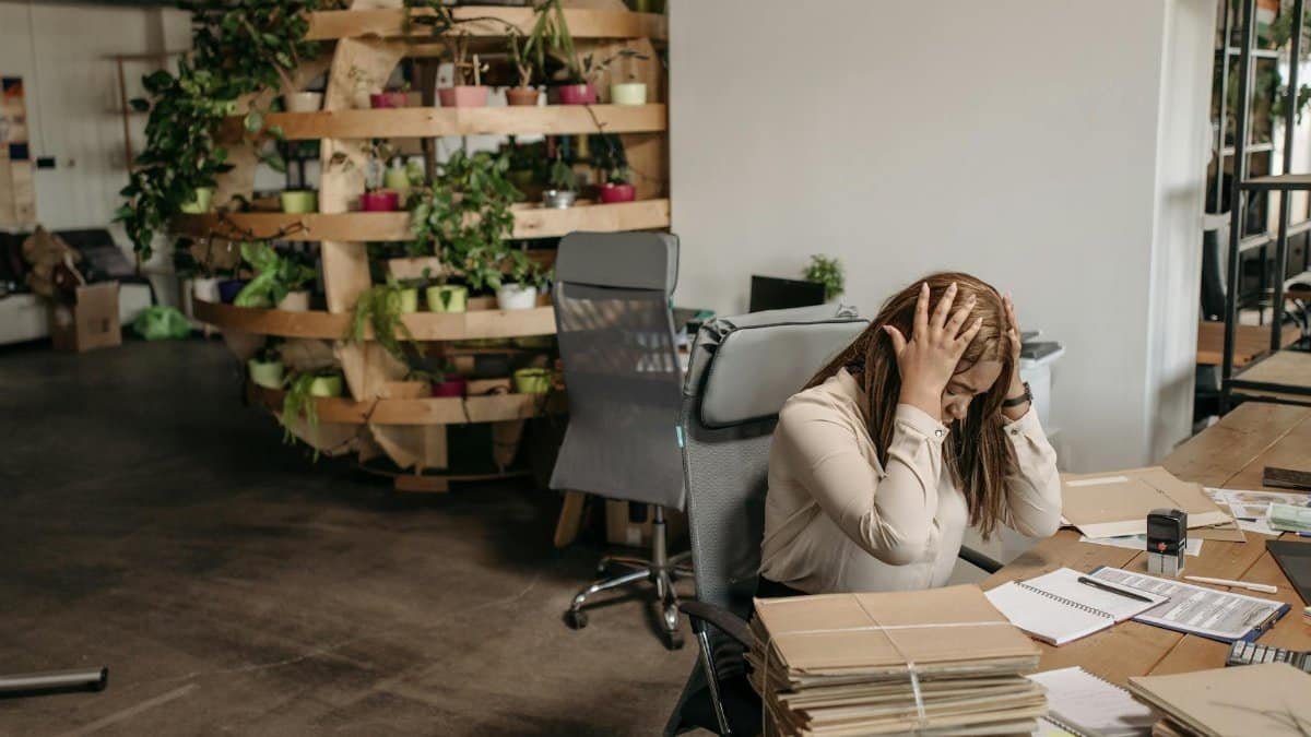 A stressed woman sits overwhelmed at her desk, surrounded by paperwork in a modern office setting.