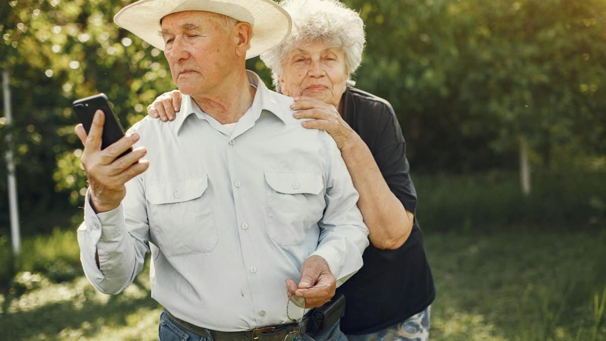 Senior couple embracing outdoors, husband using smartphone.