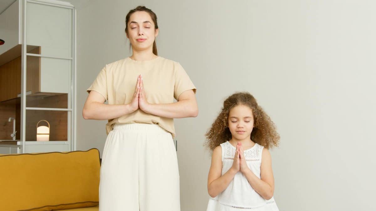 A mother and daughter practicing meditation and prayer indoors with eyes closed.