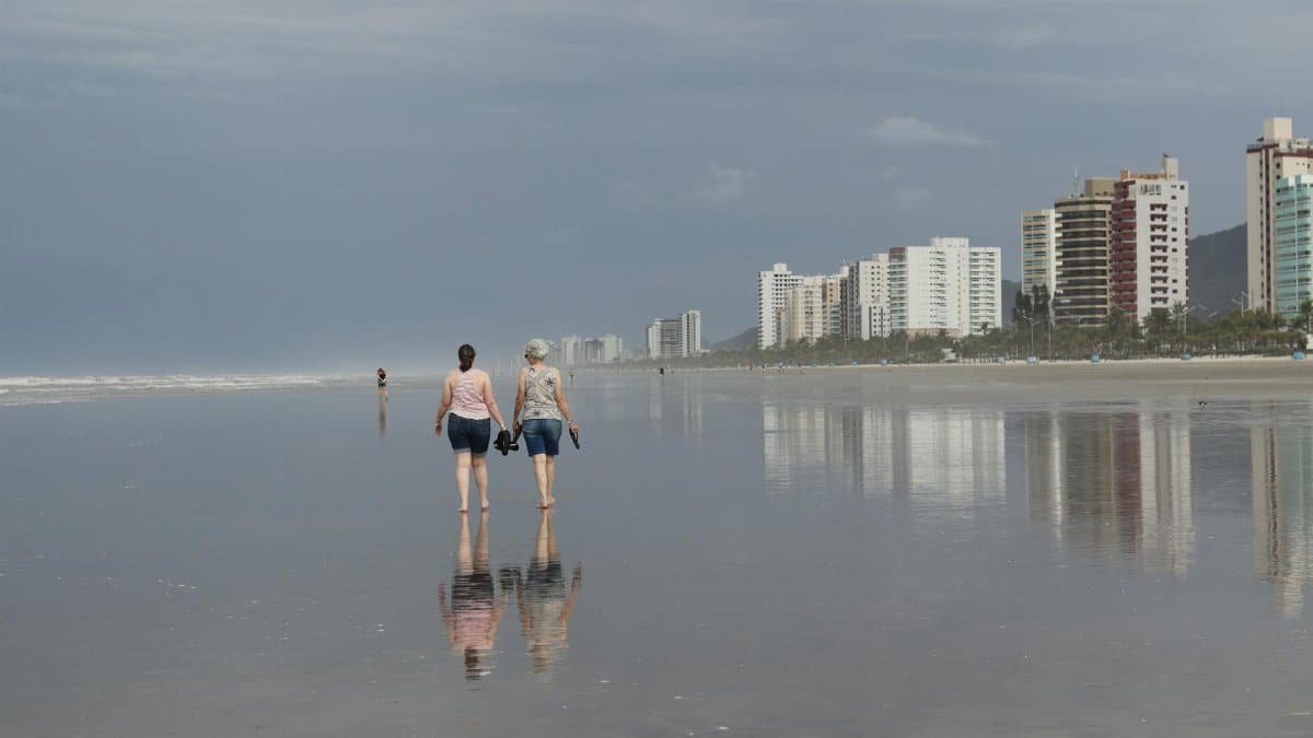 Two women walking on a serene beach with reflections, cityscape in the background.
