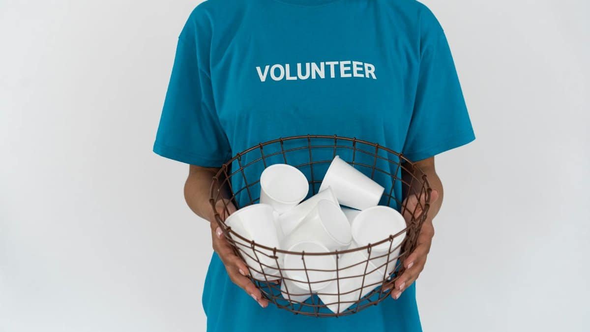 A volunteer in a blue shirt holding a wire basket with disposable cups, promoting recycling.