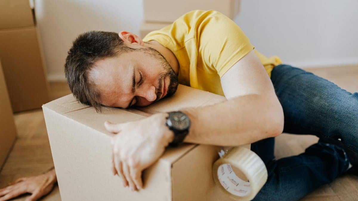 Man asleep on a cardboard box during a move, reflecting fatigue and stress.