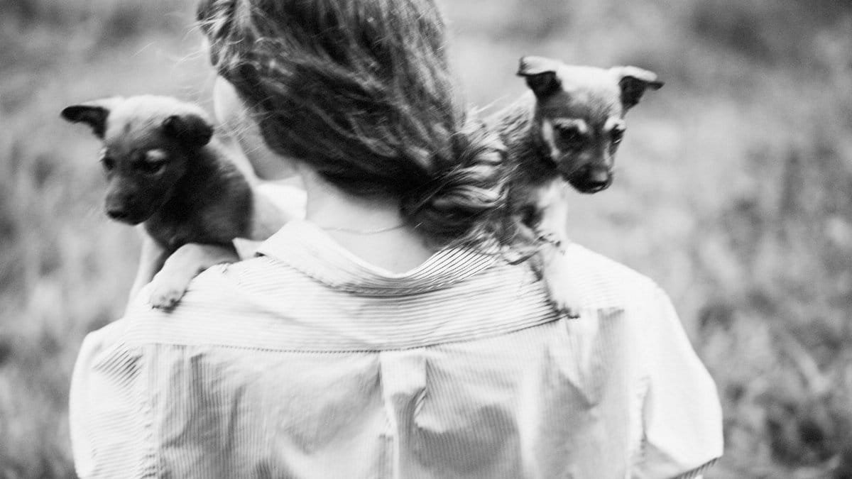 A woman holds two puppies on her shoulders in a candid, artistic black and white photograph.