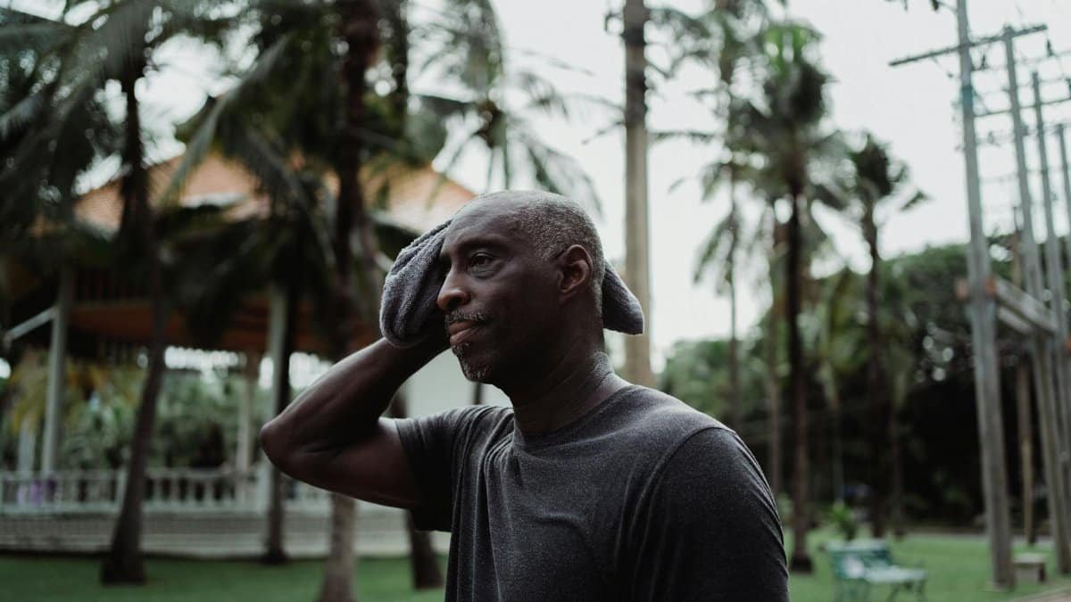 A man wiping sweat with a face towel outdoors in a tropical environment, appearing thoughtful and serious.