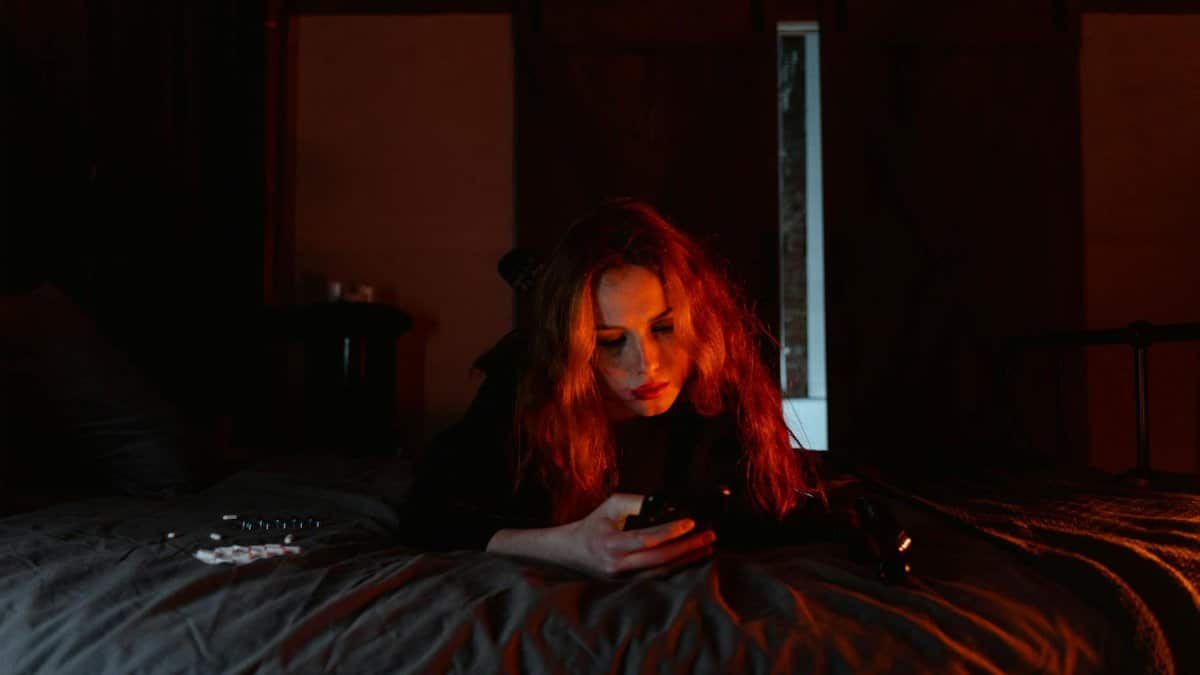 A young woman lying on a bed in a dark room, engrossed in her cellphone.