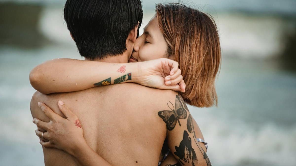 A loving couple warmly embracing at the beach, showcasing tattooed art and affection in a serene seaside setting.