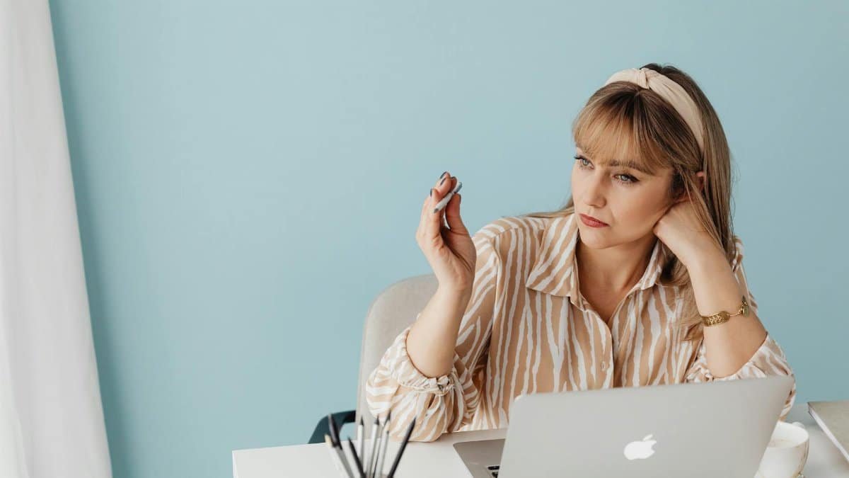 A woman relaxing in a home office setting, sitting with a laptop, fidgeting with a pencil, and enjoying a cup of coffee.