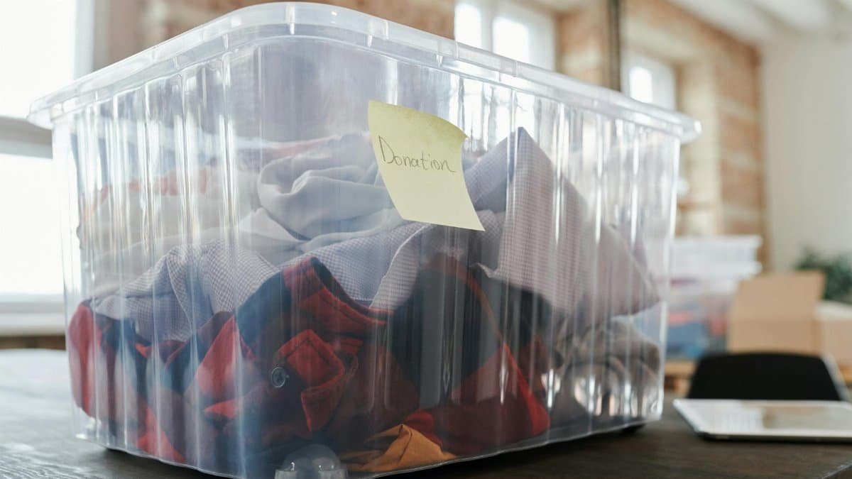 Clear plastic box filled with clothing for donation on a wooden table indoors, tagged donation.