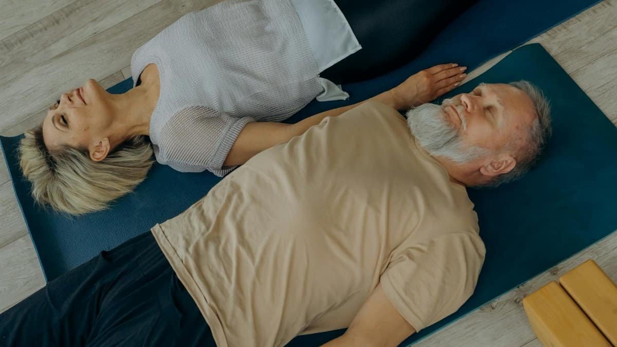 Elderly couple lying on yoga mats in a peaceful indoor setting, embracing togetherness.