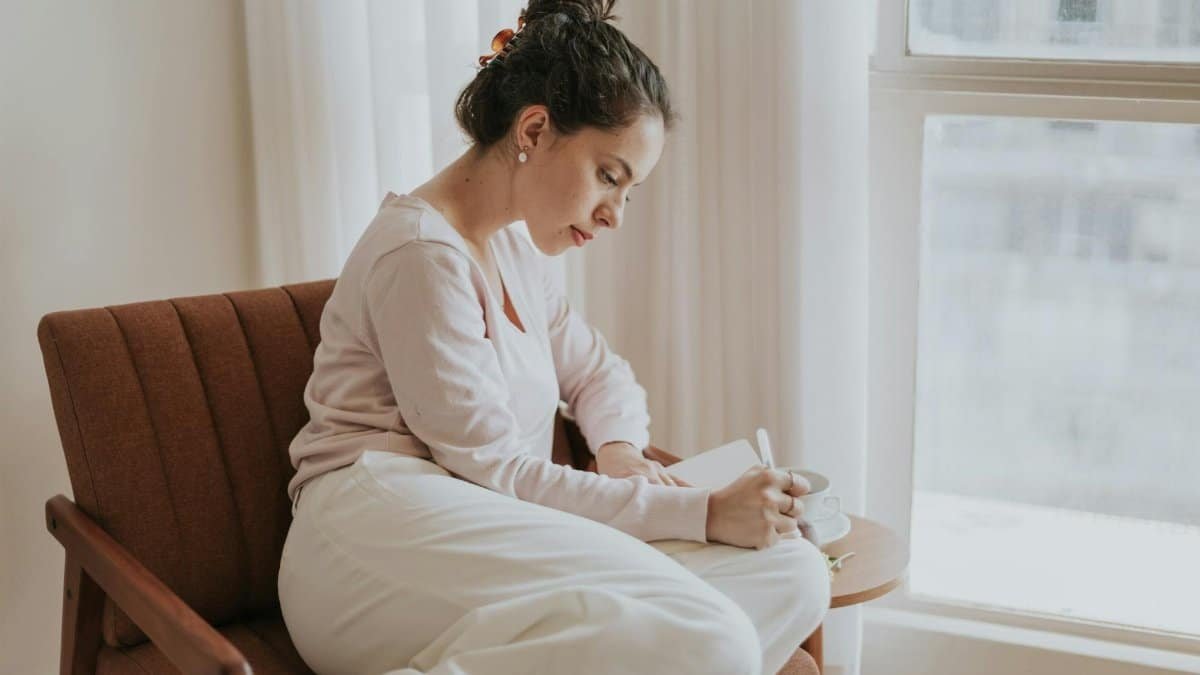 A woman comfortably journaling by a window, surrounded by soft natural light, in a cozy indoor setting.