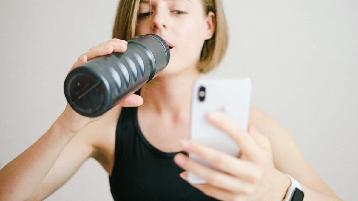 Woman drinking water while using a smartphone in a home fitness setting, highlighting modern technology and hydration.