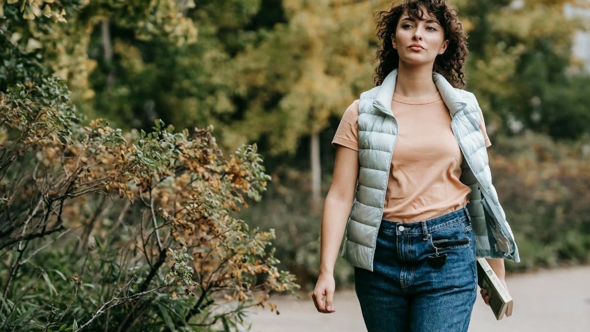 Pensive woman in casual clothes walking alone on walkway near green trees placed in city park and looking away in daytime